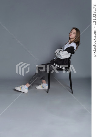 A girl sits with one arm resting on a chair, looking straight ahead. This portrait captures the spirit of modern youth, blending calm self-assurance with effortless style. A girl sits with one arm resting on a chair, looking straight ahead. This portrait captures the spirit of modern youth, blending calm self-assurance with effortless style. 121328178