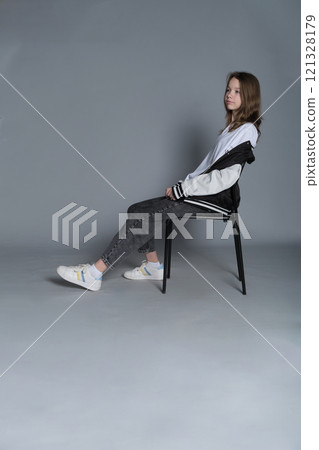 The young girl sits comfortably on a chair, her relaxed pose exuding quiet confidence. This image highlights the simplicity and authenticity of youthful moments in contemporary lifestyle. 121328179