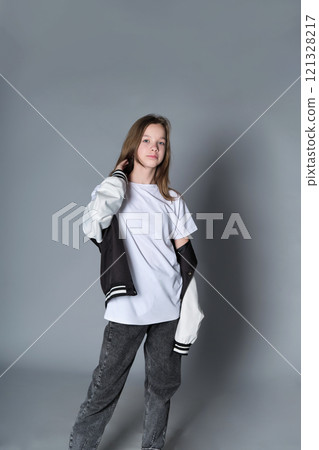 A teenage girl strikes a casual pose, wearing jeans and a varsity jacket over a white shirt. Her outfit represents youthful confidence and the freedom of self-expression in everyday wear. A teenage girl strikes a casual pose, wearing jeans and a varsity jacket over a white shirt. Her outfit represents youthful confidence and the freedom of self-expression in everyday wear. 121328217
