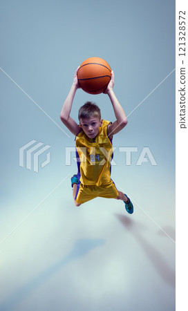 Aerial view of sportive, junior basketball athlete jumping in motion and throw ball into basket against grey studio background. Aerial view of sportive, junior basketball athlete jumping in motion and throw ball into basket against grey studio background. 121328572