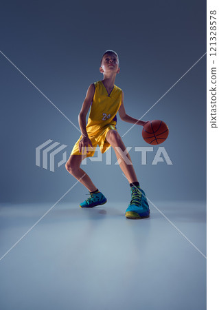 Low angle view of teenage boy, dressed vibrant basketball gear, training dribbling technique in action against grey studio background. 121328578