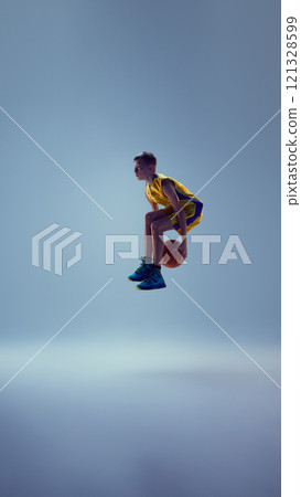 Dynamic image of little boy dressed basketball uniform, playing basketball and ready to get powerful slam dunk against grey studio background. Dynamic image of little boy dressed basketball uniform, playing basketball and ready to get powerful slam dunk against grey studio background. 121328599