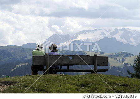 Hikers man and woman contemplating the Dolomites mountain panorama while sitting on a wooden bench 121330039
