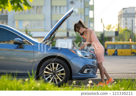 Helpless woman standing near her car with open bonnet calling road service for help. Young female driver having trouble with vehicle. Helpless woman standing near her car with open bonnet calling road service for help. Young female driver having trouble with vehicle. 121330211
