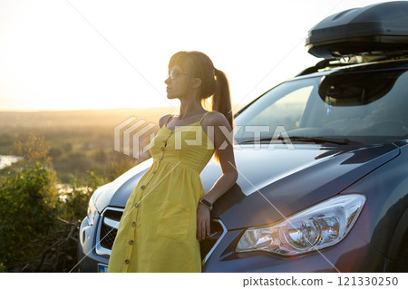 Happy young woman driver in yellow dress enjoying warm summer evening standing beside her car. Travelling and vacation concept. Happy young woman driver in yellow dress enjoying warm summer evening standing beside her car. Travelling and vacation concept. 121330250