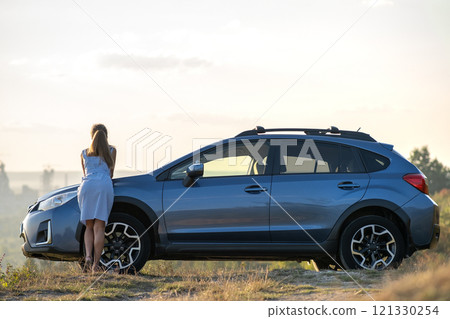 Happy woman driver in blue summer dress enjoying warm evening near her car. Travel and vacations concept. 121330254
