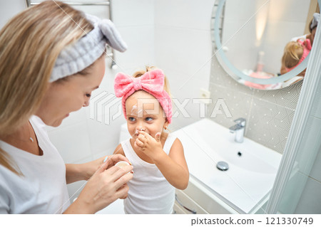 Smiling young woman and little daughter with a cosmetic bow on their heads smearing face cream on Smiling young woman and little daughter with a cosmetic bow on their heads smearing face cream on 121330749