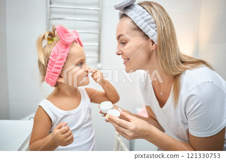 Smiling young woman and little daughter with a cosmetic bow on their heads smearing face cream on Smiling young woman and little daughter with a cosmetic bow on their heads smearing face cream on 121330753