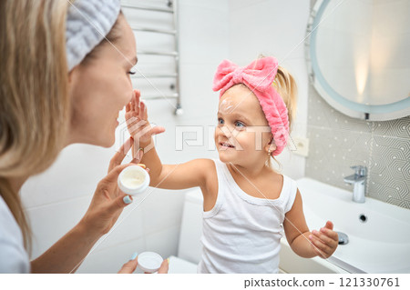 Smiling young woman and little daughter with a cosmetic bow on their heads smearing face cream on Smiling young woman and little daughter with a cosmetic bow on their heads smearing face cream on 121330761