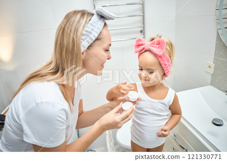 Smiling young woman and little daughter with a cosmetic bow on their heads smearing face cream on 121330771