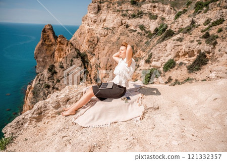 A woman is sitting on a blanket on a rocky beach, looking out at the ocean. She is wearing a white shirt and black shorts. 121332357