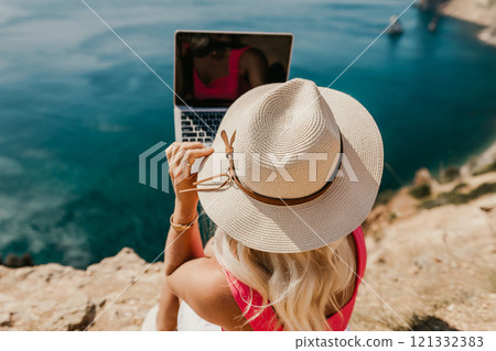 A woman is sitting on a rocky beach with a laptop in front of her. She is wearing a pink top and a straw hat. Concept of relaxation and leisure. 121332383