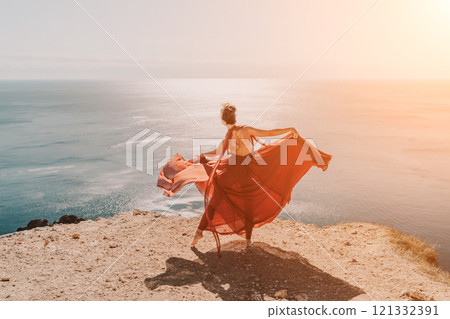 Woman red dress sea. Female dancer in a long red dress posing on a beach with rocks on sunny day. Girl on the nature on blue sky background. 121332391