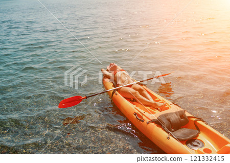 A woman is laying on a kayak in the water. The kayak is orange and has a paddle on it. The woman is wearing a red hat. 121332415