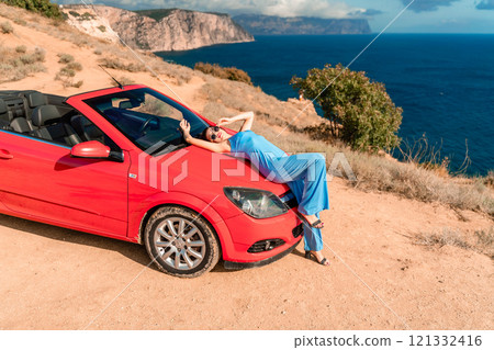 A woman is laying on top of a red convertible car. The car is parked on a dirt road near the ocean. The woman is wearing a blue dress and she is enjoying the moment. A woman is laying on top of a red convertible car. The car is parked on a dirt road near the ocean. The woman is wearing a blue dress and she is enjoying the moment. 121332416