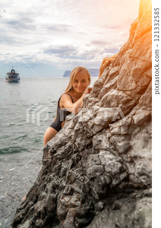 Woman swimsuit sea. Attractive blonde woman in a black swimsuit enjoying the sea air on the seashore around the rocks. Travel and vacation concept. 121332585