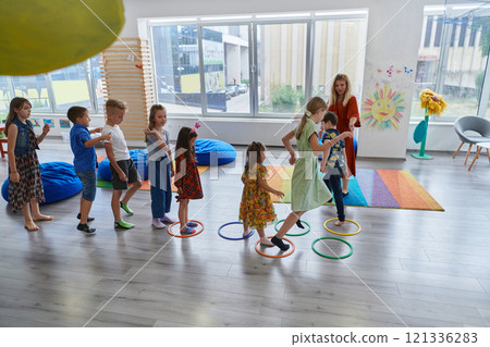 Small nursery school children with female teacher on floor indoors in classroom, doing exercise. Jumping over hula hoop circles track on the floor. Small nursery school children with female teacher on floor indoors in classroom, doing exercise. Jumping over hula hoop circles track on the floor. 121336283