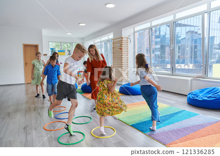 Small nursery school children with female teacher on floor indoors in classroom, doing exercise. Jumping over hula hoop circles track on the floor. Small nursery school children with female teacher on floor indoors in classroom, doing exercise. Jumping over hula hoop circles track on the floor. 121336285
