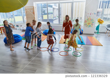Small nursery school children with female teacher on floor indoors in classroom, doing exercise. Jumping over hula hoop circles track on the floor. 121336329