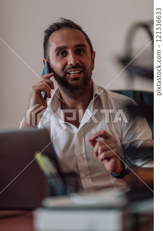 A businessman talking on his smartphone while seated in an office, showcasing his professional demeanor and active communication. A businessman talking on his smartphone while seated in an office, showcasing his professional demeanor and active communication. 121336633