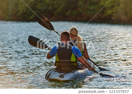 A young couple enjoying an idyllic kayak ride in the middle of a beautiful river surrounded by forest greenery in sunset time  121337057