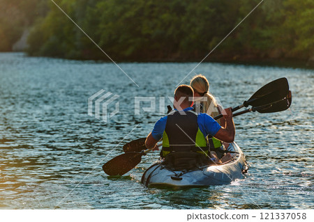 A young couple enjoying an idyllic kayak ride in the middle of a beautiful river surrounded by forest greenery in sunset time  121337058