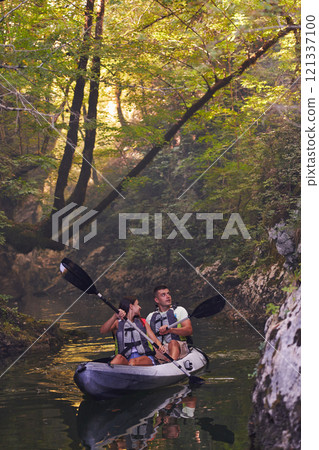A young couple enjoying an idyllic kayak ride in the middle of a beautiful river surrounded by forest greenery 121337100
