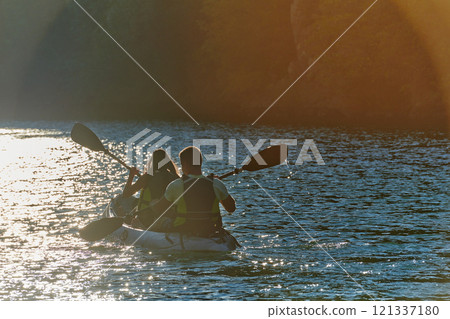 A young couple enjoying an idyllic kayak ride in the middle of a beautiful river surrounded by forest greenery in sunset time  121337180