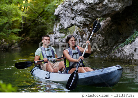 A young couple enjoying an idyllic kayak ride in the middle of a beautiful river surrounded by forest greenery 121337182