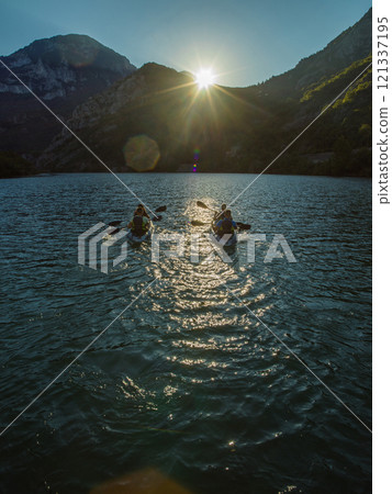 A group of friends enjoying fun and kayaking exploring the calm river, surrounding forest and large natural river canyons during an idyllic sunset. A group of friends enjoying fun and kayaking exploring the calm river, surrounding forest and large natural river canyons during an idyllic sunset. 121337195