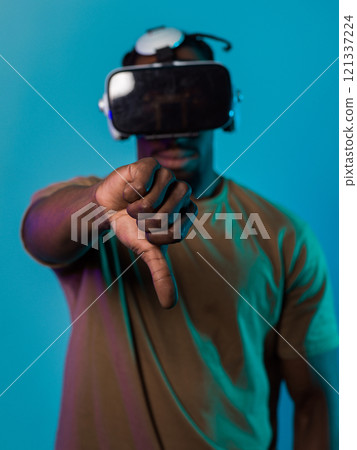 African American man, wearing virtual reality glasses, expresses dissatisfaction by pointing down with his hand while standing isolated against a blue background, portraying a modern critique within African American man, wearing virtual reality glasses, expresses dissatisfaction by pointing down with his hand while standing isolated against a blue background, portraying a modern critique within 121337224