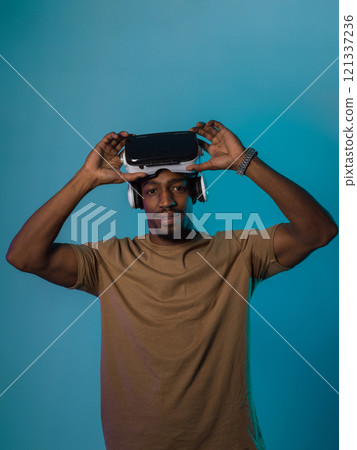 In a futuristic visual, an African American man stands isolated against a striking blue backdrop, adorned with VR glasses that transport him into a cutting-edge virtual reality experience, merging In a futuristic visual, an African American man stands isolated against a striking blue backdrop, adorned with VR glasses that transport him into a cutting-edge virtual reality experience, merging 121337236