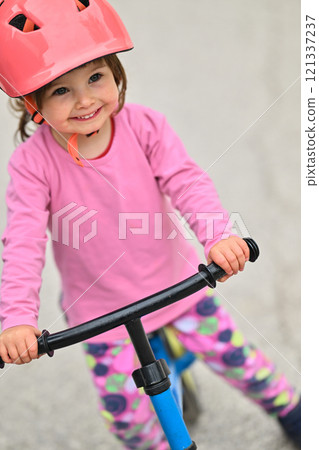 A little girl, dressed in a cute outfit and wearing a helmet, excitedly learning to ride her bike for the first time, filled with joy and determination. 121337237
