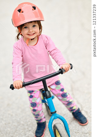 A little girl, dressed in a cute outfit and wearing a helmet, excitedly learning to ride her bike for the first time, filled with joy and determination. A little girl, dressed in a cute outfit and wearing a helmet, excitedly learning to ride her bike for the first time, filled with joy and determination. 121337239