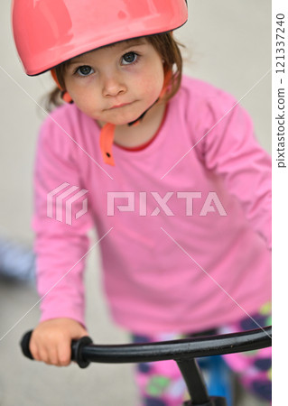 A little girl, dressed in a cute outfit and wearing a helmet, excitedly learning to ride her bike for the first time, filled with joy and determination. 121337240