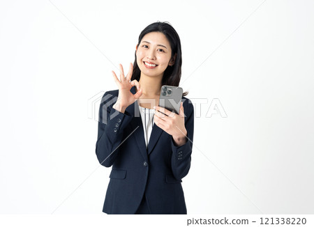 一位身穿西裝的年輕女子拿著智慧型手機，用手指做出「OK」的手勢 121338220