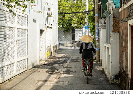 A woman wearing a straw hat rides a bicycle down a narrow street 121338828