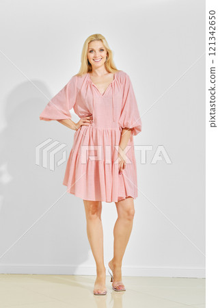 Woman with blonde hair wearing loose pink summer dress and sandals standing over white background 121342650