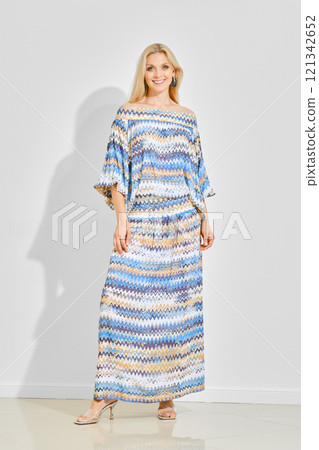 Smiling woman in a colourful boho maxi dress standing in bright room over white wall 121342652