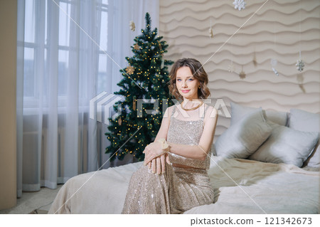 Elegant woman in glittering dress poses in front of decorated Christmas tree indoors 121342673