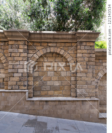 Textured stone wall featuring multiple arches creates a tranquil atmosphere in outdoor area surrounded by lush greenery 121343534