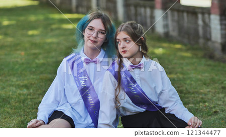 Two female graduates wearing sashes and bow ties are sitting on grass, enjoying their last school day Two female graduates wearing sashes and bow ties are sitting on grass, enjoying their last school day 121344477
