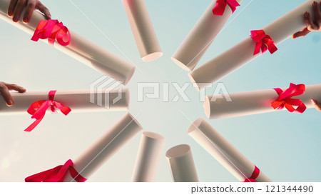 Graduates marking their achievement with red ribbon tied diplomas, forming a circle under a clear blue sky Graduates marking their achievement with red ribbon tied diplomas, forming a circle under a clear blue sky 121344490
