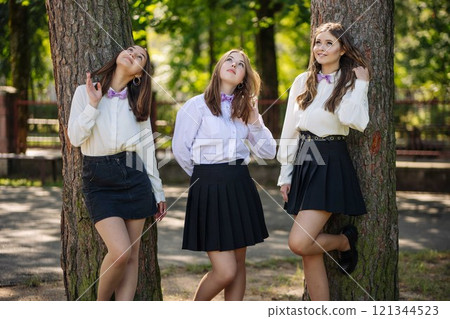 Three young girls enjoying their first day back at school, leaning against trees in a park and looking up Three young girls enjoying their first day back at school, leaning against trees in a park and looking up 121344523
