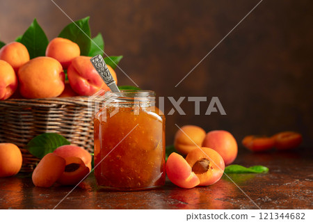 Apricot jam in glass jar and fresh fruits on a brown table. 121344682