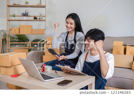 Happy Couple Celebrating Successful Online Sales with Laptops and Tablets in Modern Home Office 121346265