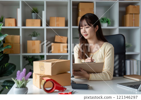 Young Female Entrepreneur Managing Online Sales in a Home Office Surrounded by Shipping Boxes and Supplies 121346297