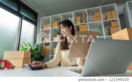 Young Woman Taking Online Sales Order in Modern Office with Laptop and Calculator Surrounded by Shipping Boxes 121346320