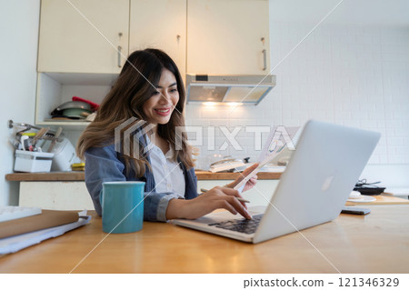 Young Freelance Woman Working from Home in the Kitchen with Laptop and Coffee, Smiling and Reviewing Documents 121346329