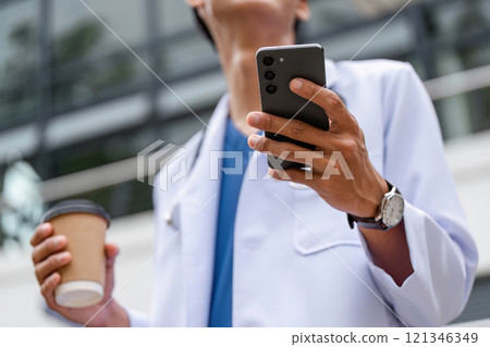 Male Doctor in Professional Uniform Holding Coffee and Smartphone Outdoors in Urban Setting 121346349